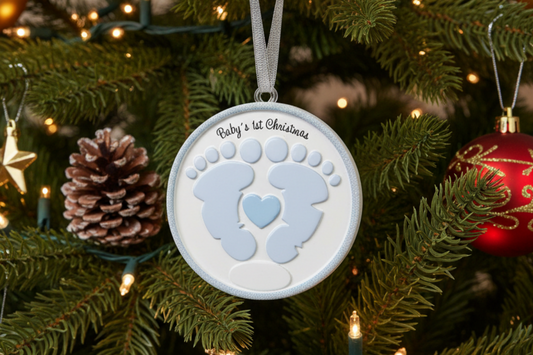 A personalized round Christmas ornament hanging on a decorated Christmas tree. The ornament is white with a light blue baby footprint design, a small light blue heart in the center, and the text 'Baby's 1st Christmas' printed at the top.