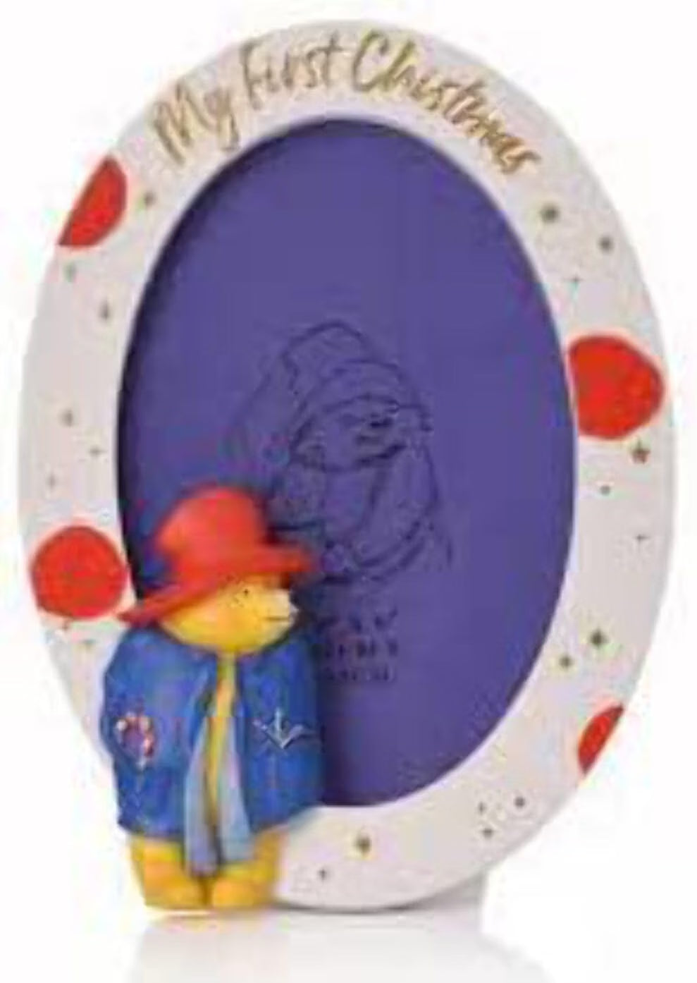 A decorative oval picture frame with a white border, red polka dots, gold script reading 'My First Christmas,' and a small standing Paddington Bear figure in front. The center insert is purple.