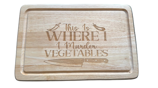 A rectangular wooden cutting board with rounded corners, featuring a laser-engraved quote that reads 'This Is WHERE I Murder VEGETABLES' .