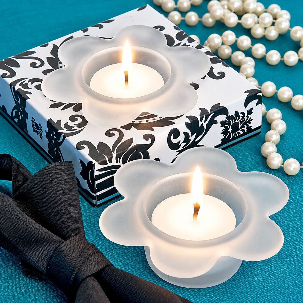 Two white flower-shaped candle holders with lit tealights, arranged on a teal surface beside a black-and-white patterned box, a black bow, and a strand of white pearls.