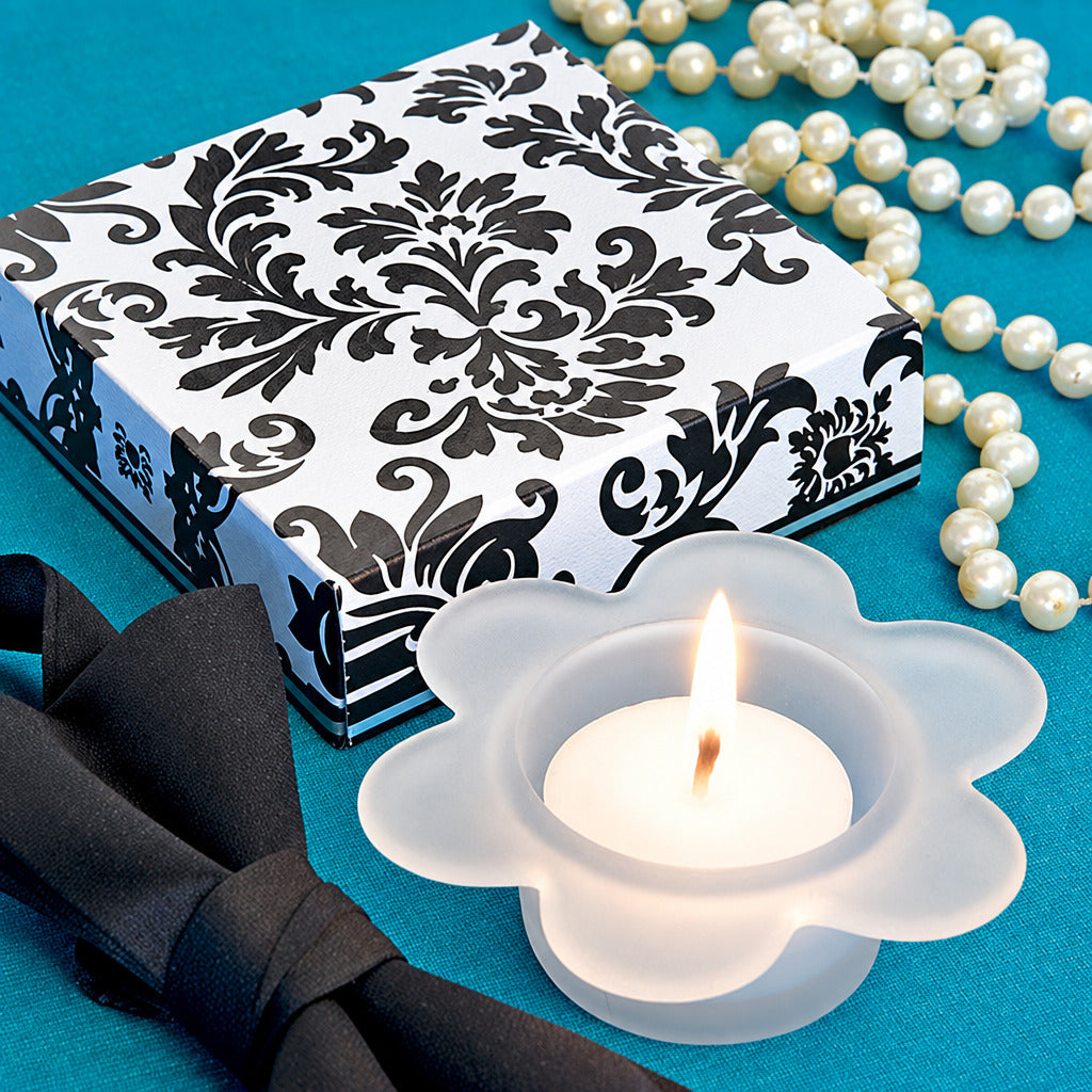 A white flower-shaped candle holders with lit tealights, arranged on a teal surface beside a black-and-white patterned box, a black bow, and a strand of white pearls.