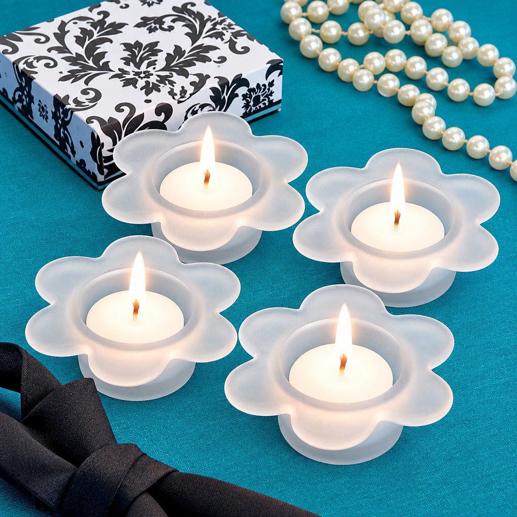 Four white flower-shaped candle holders with lit tealights, arranged on a teal surface beside a black-and-white patterned box, a black bow, and a strand of white pearls.