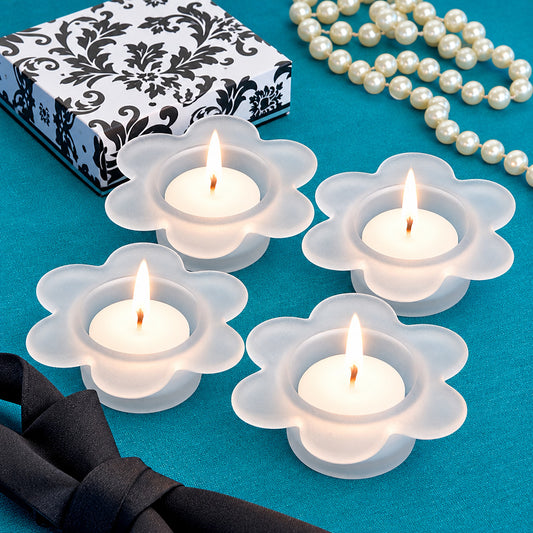 Four white flower-shaped candle holders with lit tealights, arranged on a teal surface beside a black-and-white patterned box, a black bow, and a strand of white pearls.