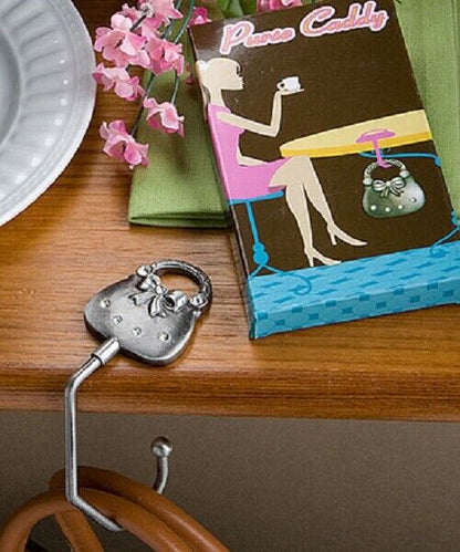 A silver metal purse caddy hook shaped like a decorative padlock, hanging on a wooden table next to a colorful 'Purse Caddy' box, a green napkin, pink flowers, and a white plate.