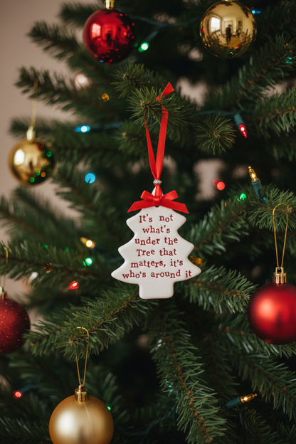 A white Christmas ornament shaped like a tree with red lettering that reads 'It's not what's under the Tree that matters, it's who's around it,' hanging from a red ribbon on a decorated Christmas tree.