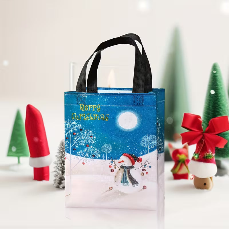 A festive Christmas gift bag with black handles, featuring a winter night scene with a snowman, a full moon, and the text 'Merry Christmas' in gold. The bag is styled on a white surface with blurred Christmas decorations in the background.