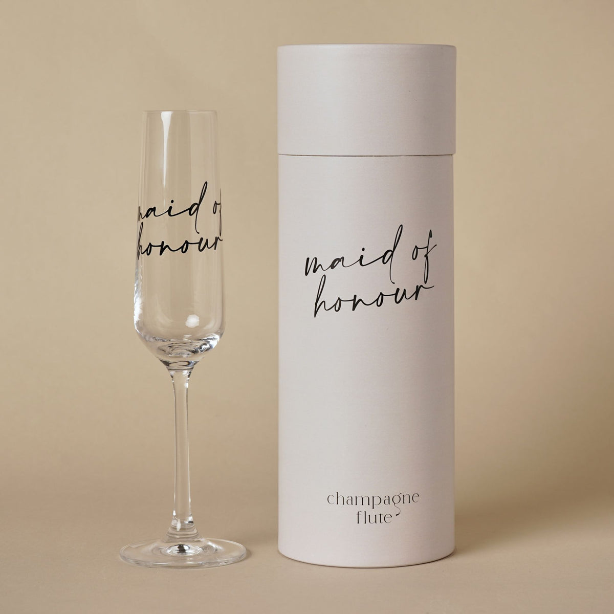 Champagne flute with “maid of honour” black script beside grey cylindrical gift tube