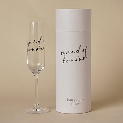 Champagne flute with “maid of honour” black script beside grey cylindrical gift tube