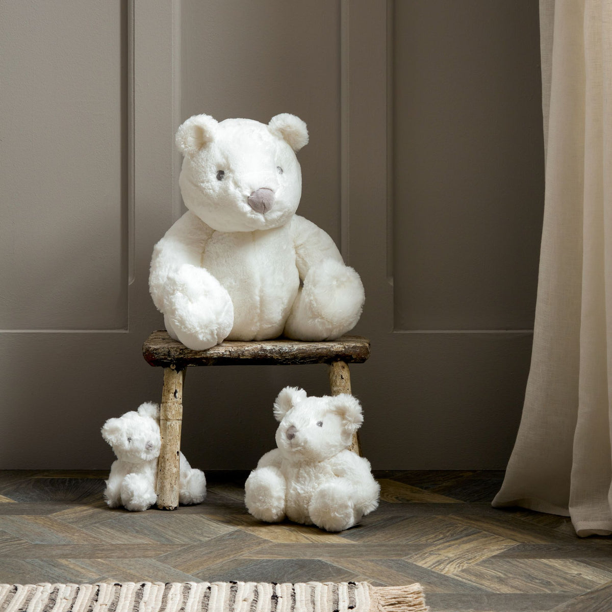 White plush bear set: one medium bear on wooden stool, two smaller bears on floor.