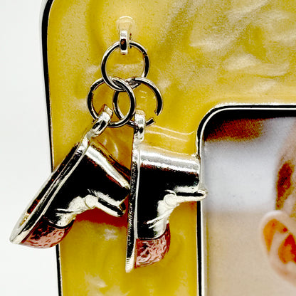 A close-up of a pair of silver-toned boot-shaped charms attached to metal rings, shown against a yellow textured background.