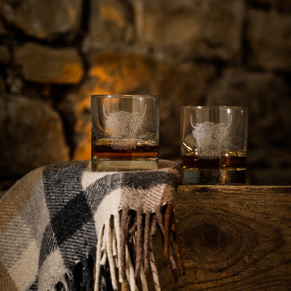 Two glasses with etched designs on a wooden surface with a stone wall background