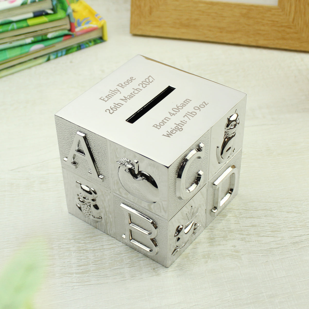 A silver metallic cube-shaped pig bank with custom engraving on one side, featuring raised letters and fruit motifs on the front, placed on a light-colored tabletop.