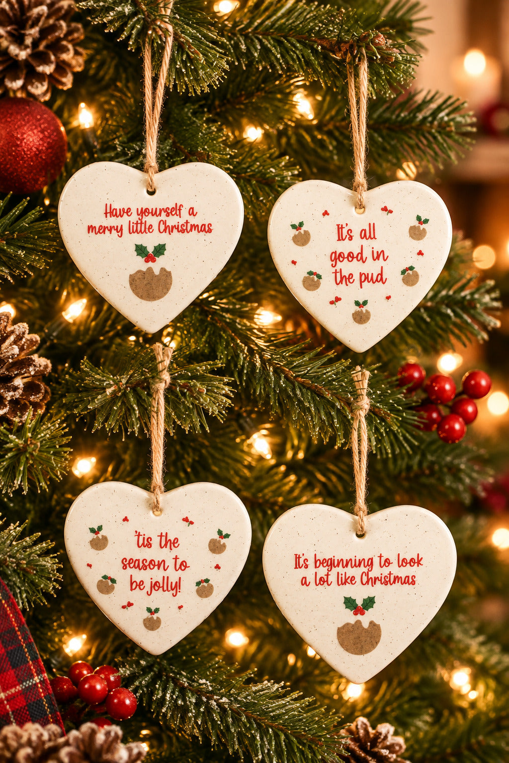 Four white heart-shaped hanging Christmas ornaments with natural twine loops, each printed with a different festive phrase in red and small brown and green holiday illustrations.