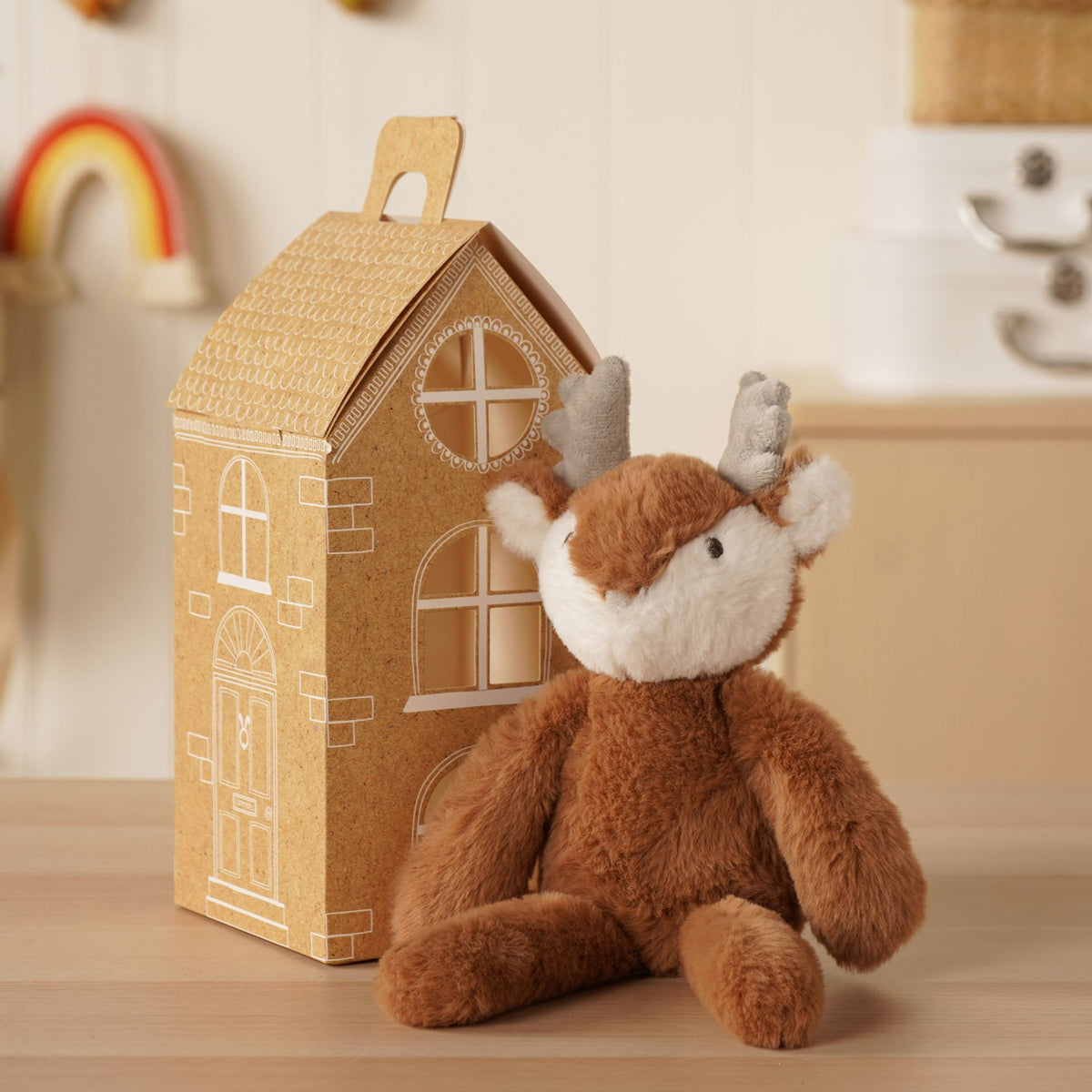 Dark brown plush long-leg reindeer sitting next to a gingerbread-style house, 30 cm