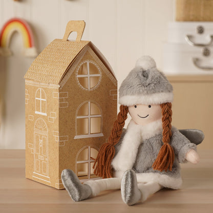 Gray plush angel doll seated with orange braids beside a decorative house