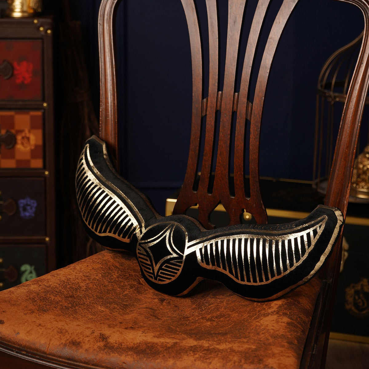 Snitch-shaped velvet cushion with gold-striped wings and central emblem on chair