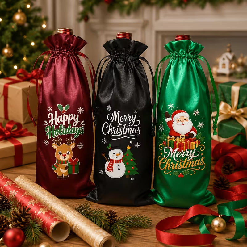 Three Christmas-themed satin bottle bags in burgundy, black, and green, decorated with festive graphics and text, displayed on a wooden table surrounded by wrapping paper, pine branches, and holiday decorations.