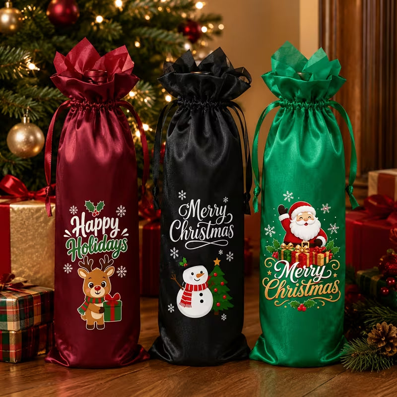 Three Christmas-themed satin drawstring gift bags in burgundy, black, and green, decorated with festive graphics and holiday greetings, displayed on a wooden table in front of a decorated Christmas tree and wrapped presents.